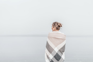 Pretty blonde woman wrapped in warm blanket standing on the cold autumn beach.