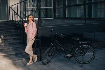 Fototapeta na wymiar Beautiful young female cyclist drinks warm coffee from a cup by the electric bicycle in the urban environment