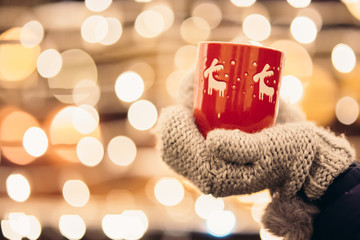 Christmas light and a woman holding in hand a red mug with hot drink - obrazy, fototapety, plakaty