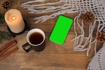 background with mug of tea, smart phone with blank green screen, candles, knitted shawl with fringe, coniferous branches and cinnamon on wood, concept winter mood, winter is love, copy space