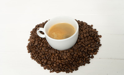 White cup with coffee in the pile of coffee beans on the white wooden background. Background photography.