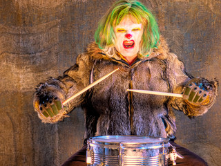 Crazy aggressive clown in wolf skin plays drum. man hates his work in his free time, dresses like clown. White face and red lips. man is ready to kill and dismember. social problem of treating madman