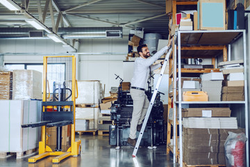 Full length of Caucasian bearded smiling graphic engineer standing on ladder and trying to reach box. Printing shop interior.