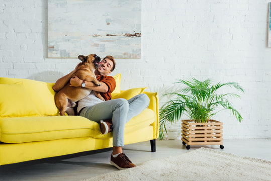 Happy man playing with french bulldog on sofa in living room