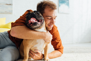 Smiling man hugging funny French bulldog on floor in living room