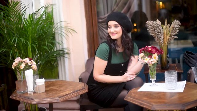 Pretty young woman in beret sitting in street cafe at table with bouquet of flowers