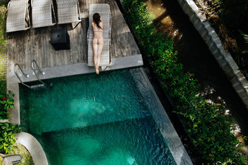 Top view of unrecognizable slim young woman in beige bikini relaxing and sunbathe near luxury swimming pool in green tropic in Ubud.Vacation concept. Drone photo