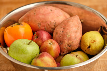 Colorful background with healthy fruits and vegetables like apples, kaki and sweet potatoes lying close together in a silver tin bowl.