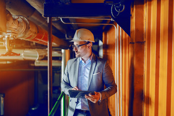 Manager in suit and with helmet on head walking around heating plant and holding tablet.
