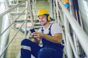 Smiling caucasian worker in overall, with helmet on head and antiphons sitting on stairs and typing message on smart phone while having pause. Factory interior.