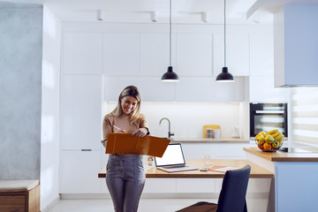 Three quarter length of caucasian blonde pretty businesswoman in sweater leaning on kitchen table and writing down statistics. On kitchen table is laptop and paperwork.