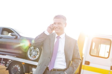 Businessman calling on smartphone for help to pick him up, his broken car is on the tow truck