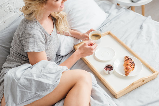 Cropped View Of Cheerful Woman Holding Cup Of Coffee Near Wooden Tray With Croissant On Bed