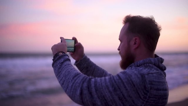 Handsome man with red beard in sweater on the beach take photo of sunset