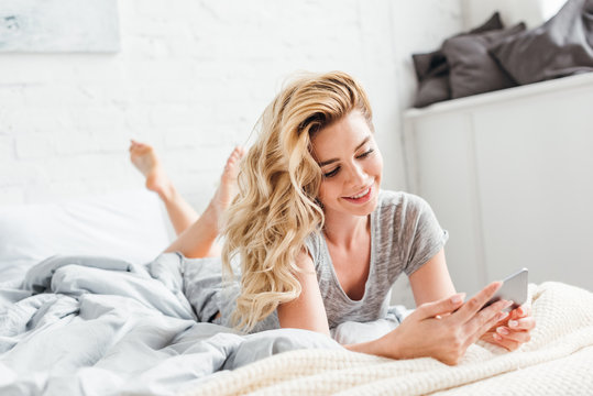 selective focus of cheerful girl using smartphone while lying on bed