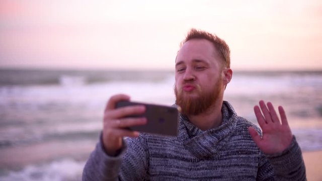 Handsome man with red beard in sweater on the beach take selfie with duck face at sunset