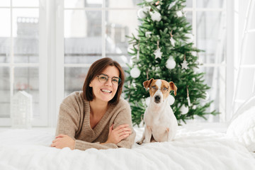 Joyful woman with short dark hair wears spectacles and round glasses, funny dog in spectacles poses near host, lie on bed, have good rest during Christmas eve, have fun together. New Year decoration
