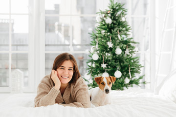 Relaxed beautiful dark haired young woman poses on bed with favourite pet, enjoys bedding time, has lazy day during winter holidays, decorated New Year tree behind, big windows. Cozy atmosphere
