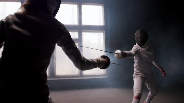 Two young women having training in a fencing duel in the smoky studio