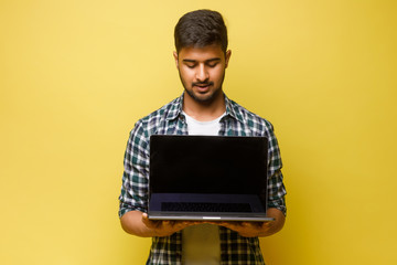 Young handsome man standing against textured yellow wall with copy space for ads, holding laptop and watching media with happy smile, sharing web content.