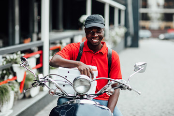 Portrait of young african guy in motorbike holding boxes with pizza and sit on his bike. Fast food...