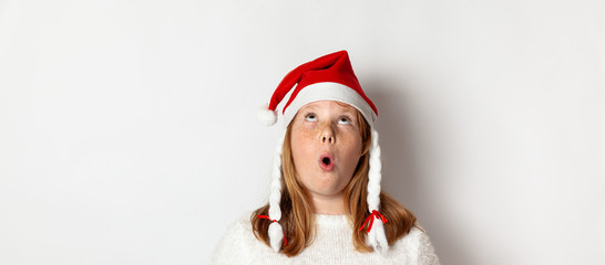 Portrait of a chubby teenager girl in a hat of Satan Claus opened her mouth in surprise and looks up, isolated on a white background.