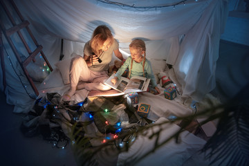 Obraz na płótnie Canvas Mother and daughter sitting in a teepee, reading stories with the flashlight in dark room with toys and pillows. Caucasian models. Home comfort, family, love, Christmas holidays, storytelling time.