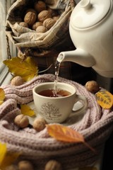 On the windowsill, boiling water is poured from the kettle into a cup of tea among walnuts, yellow leaves and a scarf