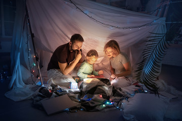 Caucasian family sitting in a teepee, reading stories with the flashlight in dark room with toys and pillows. Caucasian models. Home comfort, family, love, Christmas holidays, storytelling time. - obrazy, fototapety, plakaty