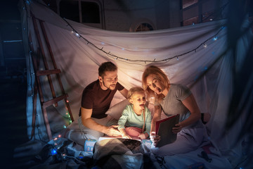 Obraz na płótnie Canvas Caucasian family sitting in a teepee, having fun and playing with the flashlight in dark room with toys and pillows. Look happy. Home comfort, family, love, Christmas holidays, storytelling time.