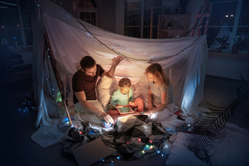 Obraz na płótnie Canvas Caucasian family sitting in a teepee, having fun and playing with the flashlight in dark room with toys and pillows. Look happy. Home comfort, family, love, Christmas holidays, storytelling time.