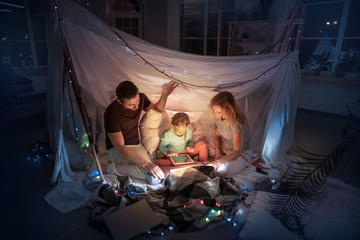 Caucasian family sitting in a teepee, having fun and playing with the flashlight in dark room with...
