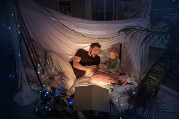 Obraz na płótnie Canvas Father and daughter sitting in a teepee, having fun, playing with the flashlight in dark room with toys and pillows. Look happy. Home comfort, family, love, Christmas holidays, storytelling time.