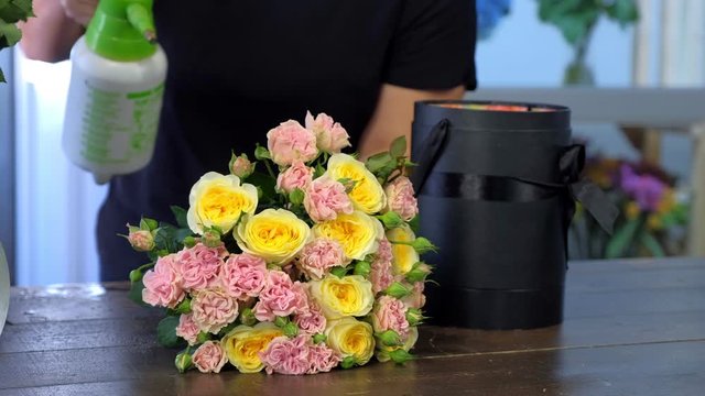 Professional florist woman spraying flowers with water making bouquet of roses putting it in packaging box, closeup hands. Floral business concept. Creates bunch composition in floristic studio store.