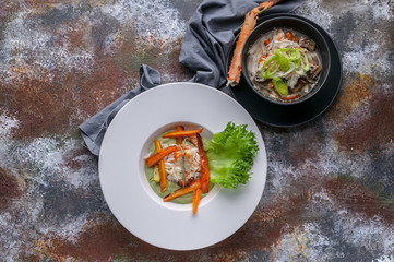 Close-up salad with snow crab meat, soup with snow crab meat, udon noodles and vegetables. Serving on a gray napkin and a rusty background. Top view