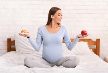 Pregnant Lady Choosing Between Apple And Croissants Sitting In Bedroom