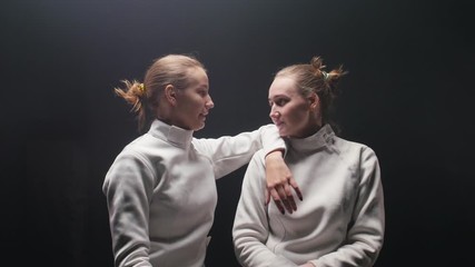 Two young women fencers in white protective suits talking to each other - leaning on the shoulder of the friend