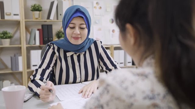 asian islam woman consultant talking with lady client in bright modern office. arabian female financial advisor explain paperwork and documents to client at meeting. friendly smiling malay girl sales