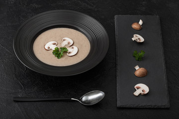 Black restaurant plate of creamy chestnut champignon mushroom soup on black table background with black stone board and fresh mushrooms and spoon.