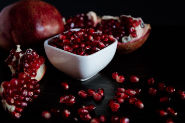 White bowl with pomegranate seeds and ripe pomegranate on dark wooden background.