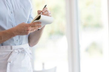 Midsection of young waiter writing on notepad while standing at restaurant