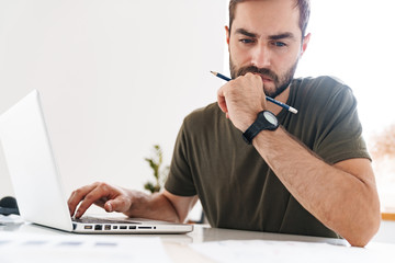 Image of young concentrated man typing on laptop and making notes