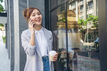A beautiful Asian woman is walking, drinking coffee, holding a cup of coffee and talking on the phone while traveling to work in the morning.