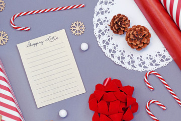 Flat lay design concept for buying Christmas presents with empty shopping list and seasonal decoration like candy canes, wooden snowflakes, gift wrapping paper and ribbon on gray background
