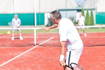 Man swinging racket while playing tennis doubles on red court during summer weekend