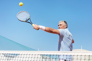 Low angle view of confident mature man hitting tennis ball with racket on court against clear blue sky
