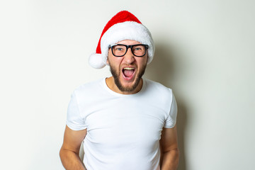 Young man in glasses and a Santa Claus hat shouts on a white background. Christmas concept. Gesture victory, super, cheers, joy