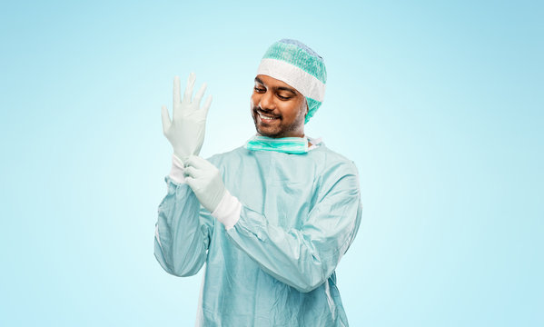 Medicine, Surgery And People Concept - Smiling Indian Male Doctor Or Surgeon In Protective Wear Putting Glove On Over Grey Background