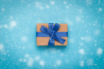 Gift box with a blue ribbon on a blue background with the effect of falling snow. Concept of gifts for Christmas and New Year. Flat lay, top view