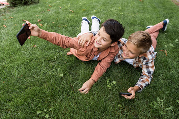 cheerful boy taking selfie on smartphone while lying on grass with brother